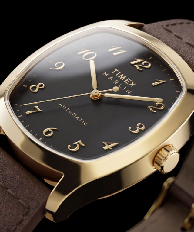 Zbliżenie na zegarek Timex Marlin Chatham Automatic w złotym kolorze, ukazujące jego czarną tarczę, nakładane cyfry arabskie w złotym kolorze, wskazówki w złotym kolorze i brązowy skórzany pasek