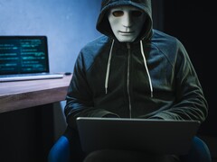 Zamaskowana, zakapturzona postać pracuje na laptopie w zaciemnionym pokoju.