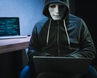 Zamaskowana, zakapturzona postać pracuje na laptopie w zaciemnionym pokoju.