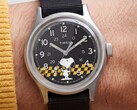 Zbliżenie na czarną tarczę zegarka Timex MK1 przedstawiającą postać Snoopy’ego na tle grafiki w żółto-czarną kratkę, z arabskimi znacznikami godzin oraz świecącymi wskazówkami.