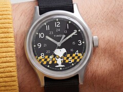 Zbliżenie na czarną tarczę zegarka Timex MK1 przedstawiającą postać Snoopy’ego na tle grafiki w żółto-czarną kratkę, z arabskimi znacznikami godzin oraz świecącymi wskazówkami.