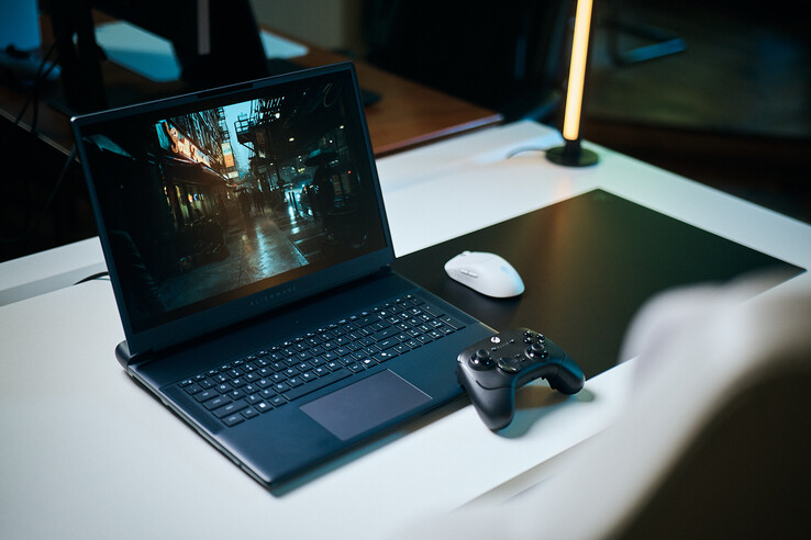 Otwarty laptop do gier marki Alienware ustawiony na białym biurku w studiu, na ekranie którego wyświetla się ciemna, odbijająca światło scena uliczna o ostrych szczegółach uzyskanej dzięki technologii śledzenia ścieżki (path tracing), w połączeniu z czarnym kontrolerem i białą myszą bezprzewodową.