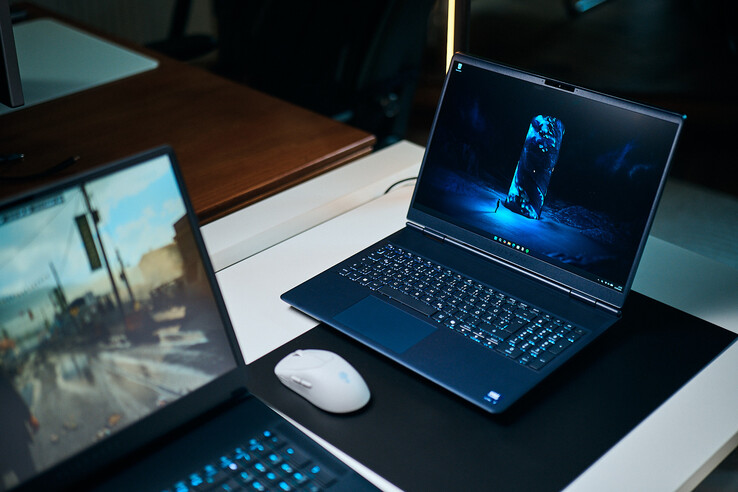 Ciemnoniebieski laptop do gier klasy średniej (Alienware Aurora 16X) ustawiony pod kątem na podkładce biurkowej z podświetlaną niebieską tapetą w stylu science fiction, umieszczony obok białej myszy bezprzewodowej, z innym laptopem zamazanym na pierwszym planie.