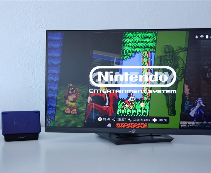 Retroid Pocket Flip 2 w kolorze fioletowym umieszczony na stacji dokującej obok monitora, z ekranem telewizora wyświetlającym interfejs emulatora retro NES. (Źródło obrazu: Retro Game Corps na YouTube)