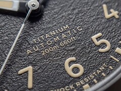 Zbliżenie na teksturowaną czarną tarczę zegarka Timex Expedition Field Post Titanium z cyframi arabskimi, świecącymi wskazówkami i znacznikami oraz okienkiem datownika.