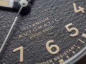 Zbliżenie na teksturowaną czarną tarczę zegarka Timex Expedition Field Post Titanium z cyframi arabskimi, świecącymi wskazówkami i znacznikami oraz okienkiem datownika.