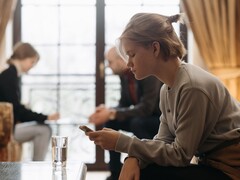 Nastolatka patrzy na smartfon, a w tle, poza ostrością, siedzą dwie dorosłe osoby.