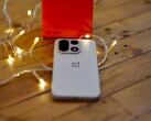 Na drewnianym stole, otoczonym ciepłymi lampkami łańcuchowymi, leży telefon OnePlus 15, a w tle widoczne jest jaskrawoczerwone opakowanie detaliczne marki OnePlus.