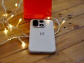 Na drewnianym stole, otoczonym ciepłymi lampkami łańcuchowymi, leży telefon OnePlus 15, a w tle widoczne jest jaskrawoczerwone opakowanie detaliczne marki OnePlus.