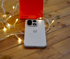 Na drewnianym stole, otoczonym ciepłymi lampkami łańcuchowymi, leży telefon OnePlus 15, a w tle widoczne jest jaskrawoczerwone opakowanie detaliczne marki OnePlus.
