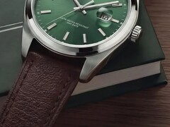 Zbliżenie zegarka Timex Legacy Day and Date, ukazujące jego zieloną tarczę ze szlifem słonecznym, lunetę w srebrnym kolorze i szczotkowane uszy koperty, żłobioną koronkę oraz fakturowany brązowy skórzany pasek na tle ciemnozielonego pudełka i drewnianej powierzchni.