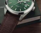 Zbliżenie zegarka Timex Legacy Day and Date, ukazujące jego zieloną tarczę ze szlifem słonecznym, lunetę w srebrnym kolorze i szczotkowane uszy koperty, żłobioną koronkę oraz fakturowany brązowy skórzany pasek na tle ciemnozielonego pudełka i drewnianej powierzchni.