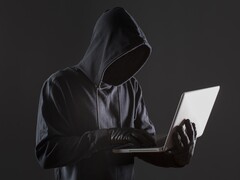 Zakapturzony haker obsługujący laptopa na ciemnym tle.