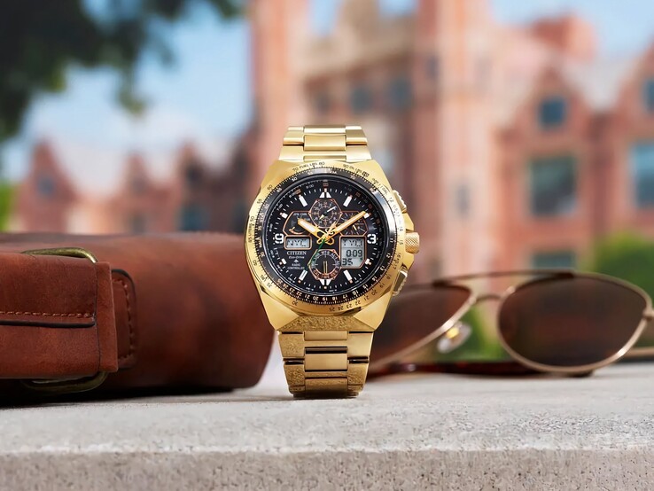Złocisty zegarek pilotażowy Citizen Promaster Skyhawk A-T JY8144-50E ustawiony pionowo na półce obok brązowej skórzanej torby i okularów przeciwsłonecznych, z zamazanym pejzażem miejskim w tle.