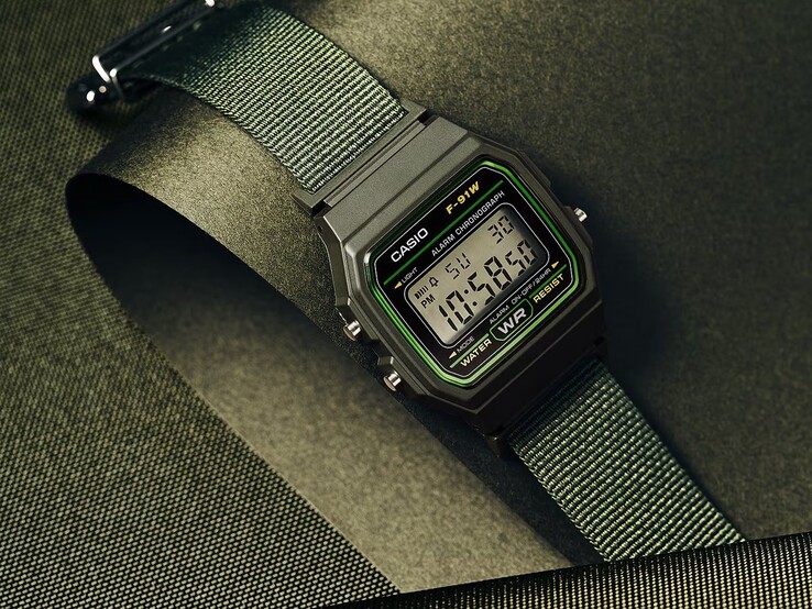 Cyfrowy zegarek Casio F-91WF-3A z czarną kopertą z zielonymi akcentami oraz zielonym paskiem z tkaniny, sfotografowany na ciemnozielonym tle o fakturze.