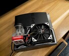 Niestandardowe chłodzenie wodne PlayStation 3 z czerwoną pompą SysCooling i dużym czarnym wentylatorem zamontowanym wewnątrz obudowy, umieszczone na drewnianym biurku. (Źródło zdjęcia: Zac Builds via YouTube)
