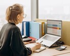 Kobieta w okularach pracuje nad arkuszem kalkulacyjnym na laptopie, a na biurku leżą segregatory i kalkulator