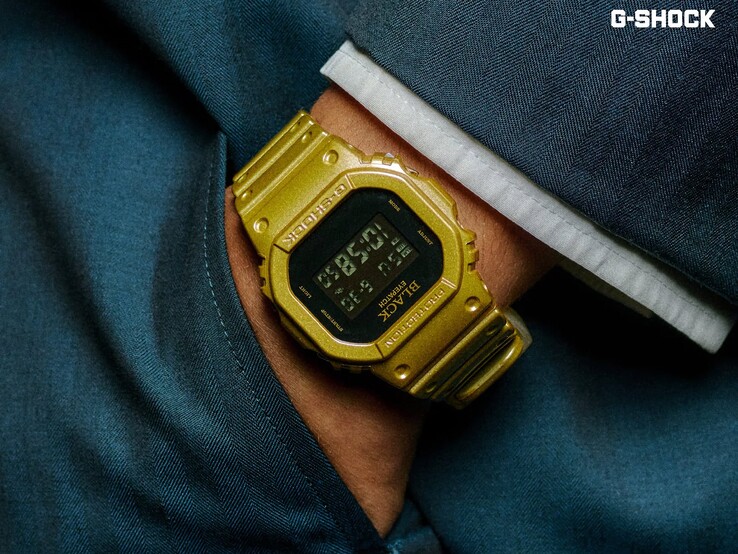 Zbliżenie na złoty zegarek Casio G-Shock DW-5600BEP-9 noszony na nadgarstku na tle ciemnoniebieskiej tkaniny. Zegarek posiada czarny wyświetlacz cyfrowy, złotą kopertę i pasek oraz logo BlackEyePatch na ramce i tarczy.