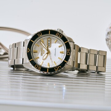 Zegarek Seiko 5 Sports SKX HDB004 ze srebrzystobiałą tarczą, czarnym obrotowym bezelem z oznaczeniami w kolorze złota, wskazówkami i indeksami w kolorze złota oraz bransoletą ze stali nierdzewnej, sfotografowany pod kątem.