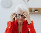 Starsza kobieta o siwych włosach, w okularach, rozmawiająca przez telefon w jasnym salonie, przyciskająca dłoń do skroni.