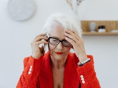 Starsza kobieta o siwych włosach, w okularach, rozmawiająca przez telefon w jasnym salonie, przyciskająca dłoń do skroni.