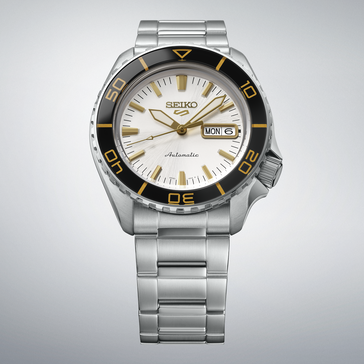 Seiko 5 Sports SKX HDB004 ze srebrno-białą tarczą, czarnym bezelem z oznaczeniami w kolorze złotym, wskazówkami i indeksami w złotym odcieniu oraz bransoletą ze stali nierdzewnej, przedstawiony na jasnoszarym tle.